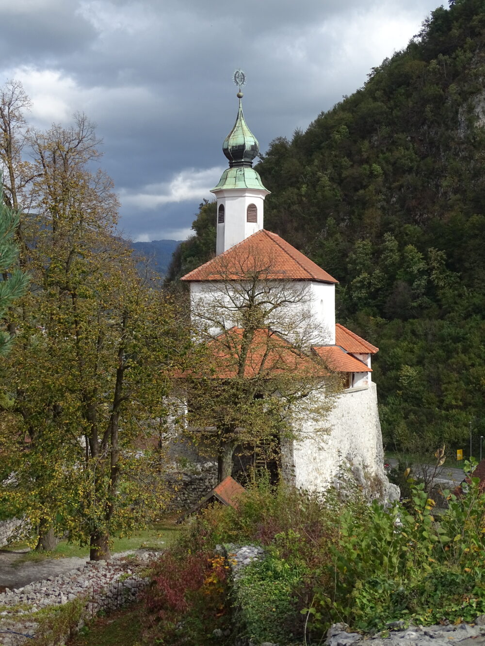 Pogled na kapelo izpred malograjskega stražnega stolpa. Zgradba je postavljena neposredno nad pečinami na jugovzhodni strani grajskega griča in povsem vpeta v obrambno obzidje gradu. V ozadju vidimo pobočja Starega gradu.