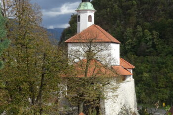 Pogled na kapelo izpred malograjskega stražnega stolpa. Zgradba je postavljena neposredno nad pečinami na jugovzhodni strani grajskega griča in povsem vpeta v obrambno obzidje gradu. V ozadju vidimo pobočja Starega gradu.